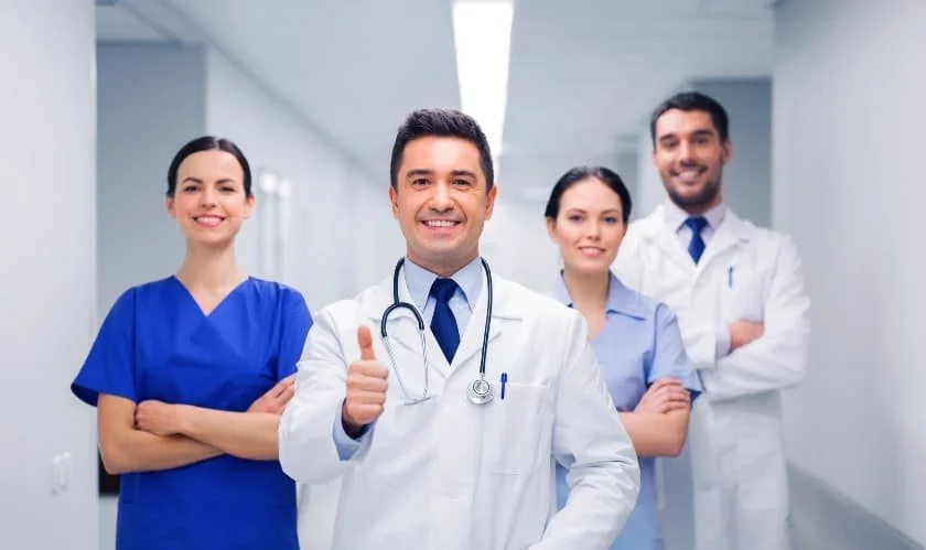 Group of four medical professionals standing in hospital corridor, smiling, with the doctor in front giving a thumbs-up.