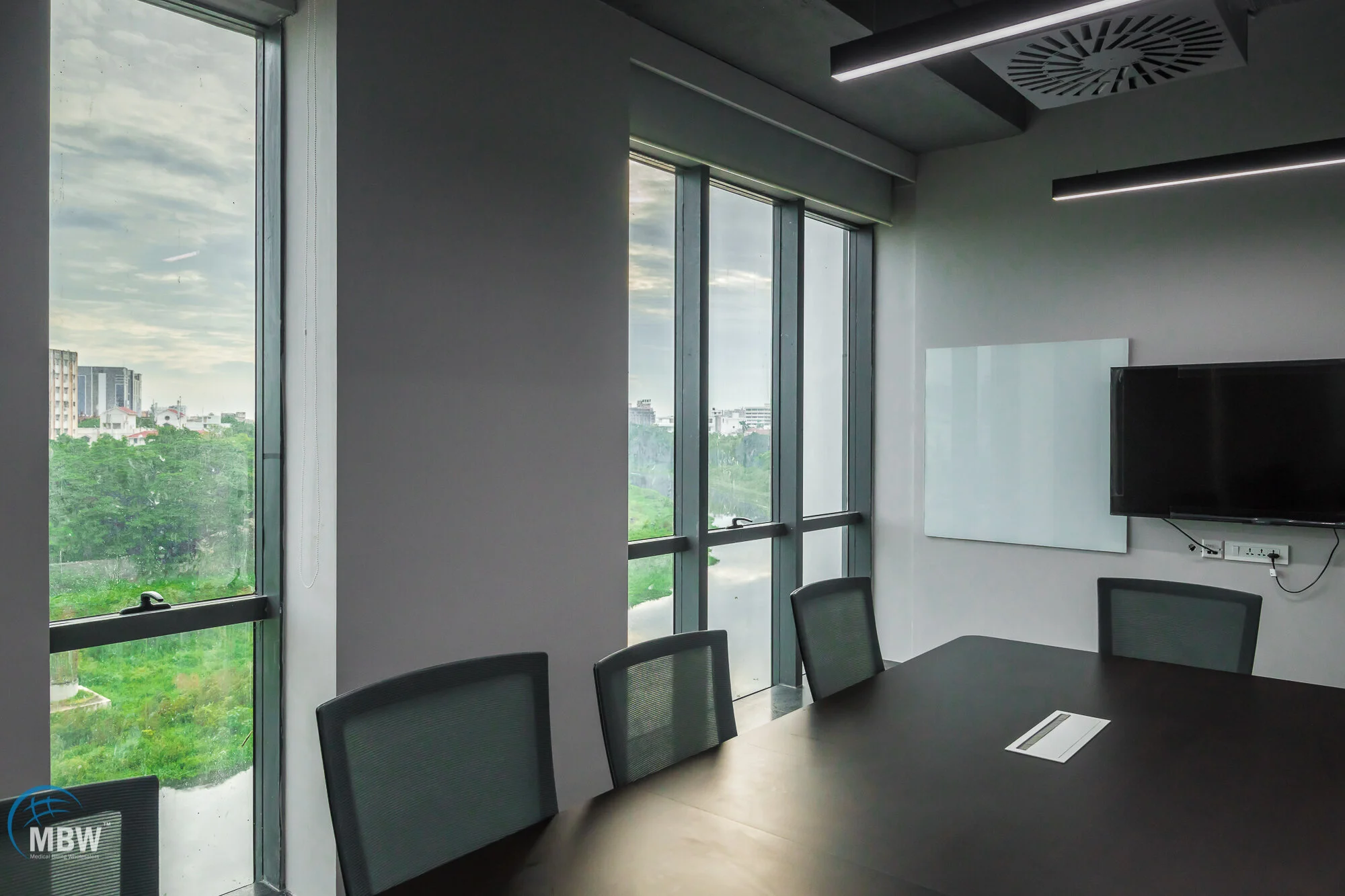 A conference room with a large black table, several chairs, large windows showing an outdoor view, a mounted flat-screen TV, and whiteboards on the wall.