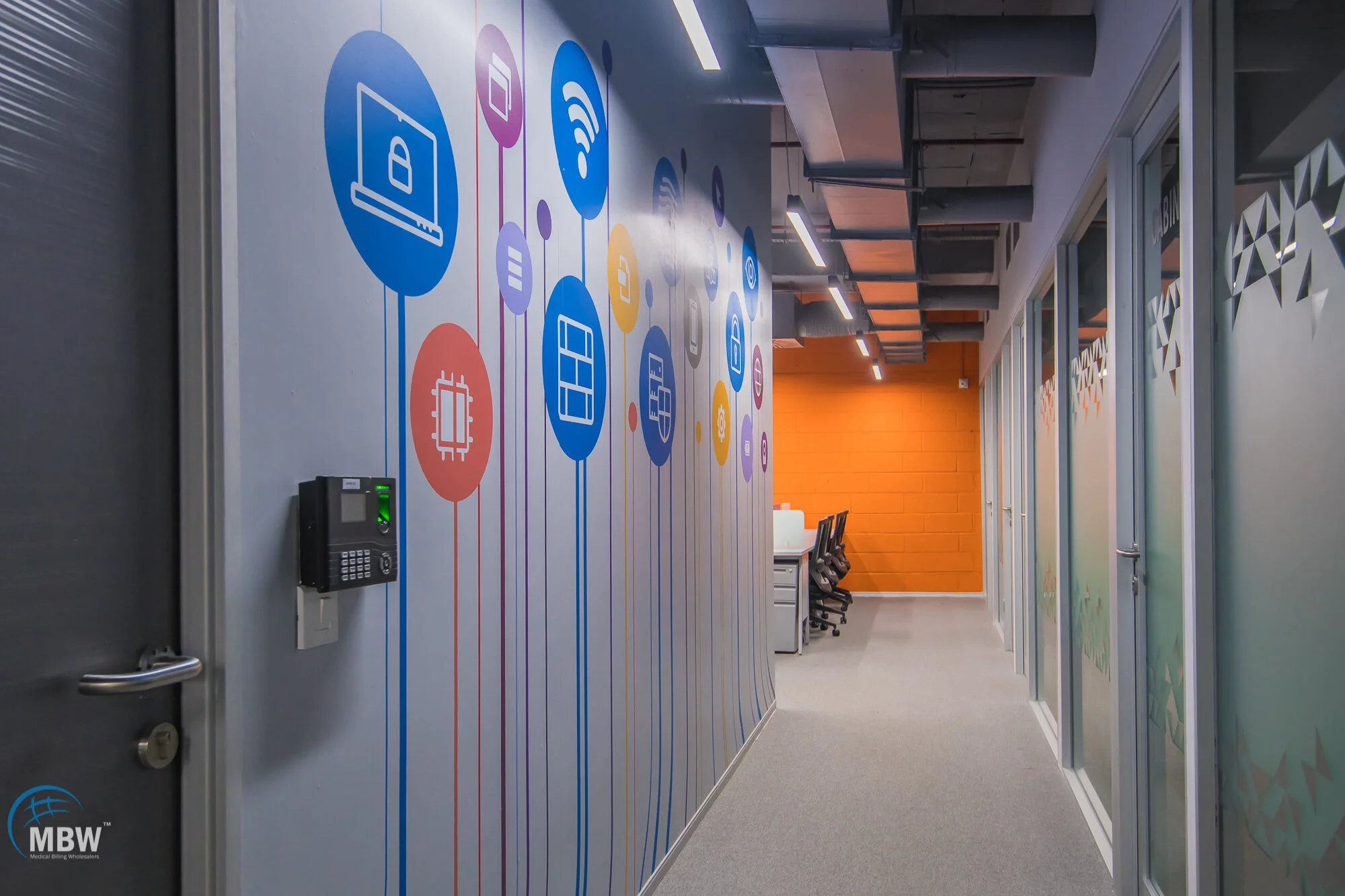 Modern office hallway with colorful wall graphics featuring technology icons, glass doors with geometric patterns, and a workspace at the end with chairs and computers.