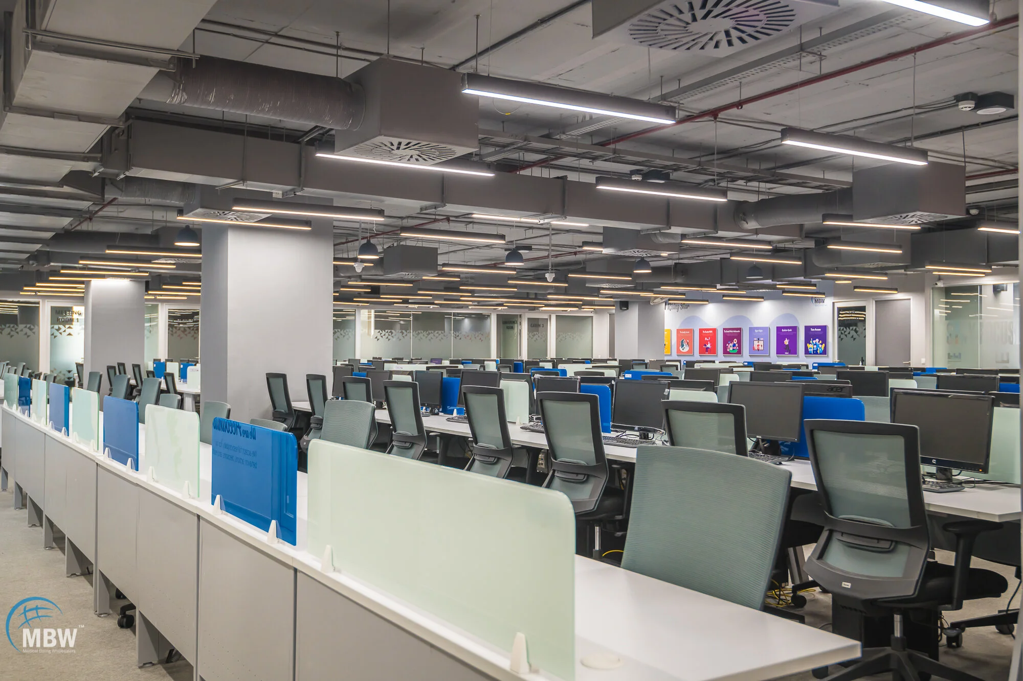 Empty call center with multiple rows of desks equipped with computer monitors, ergonomic chairs, and partition screens, illuminated by modern overhead lighting.