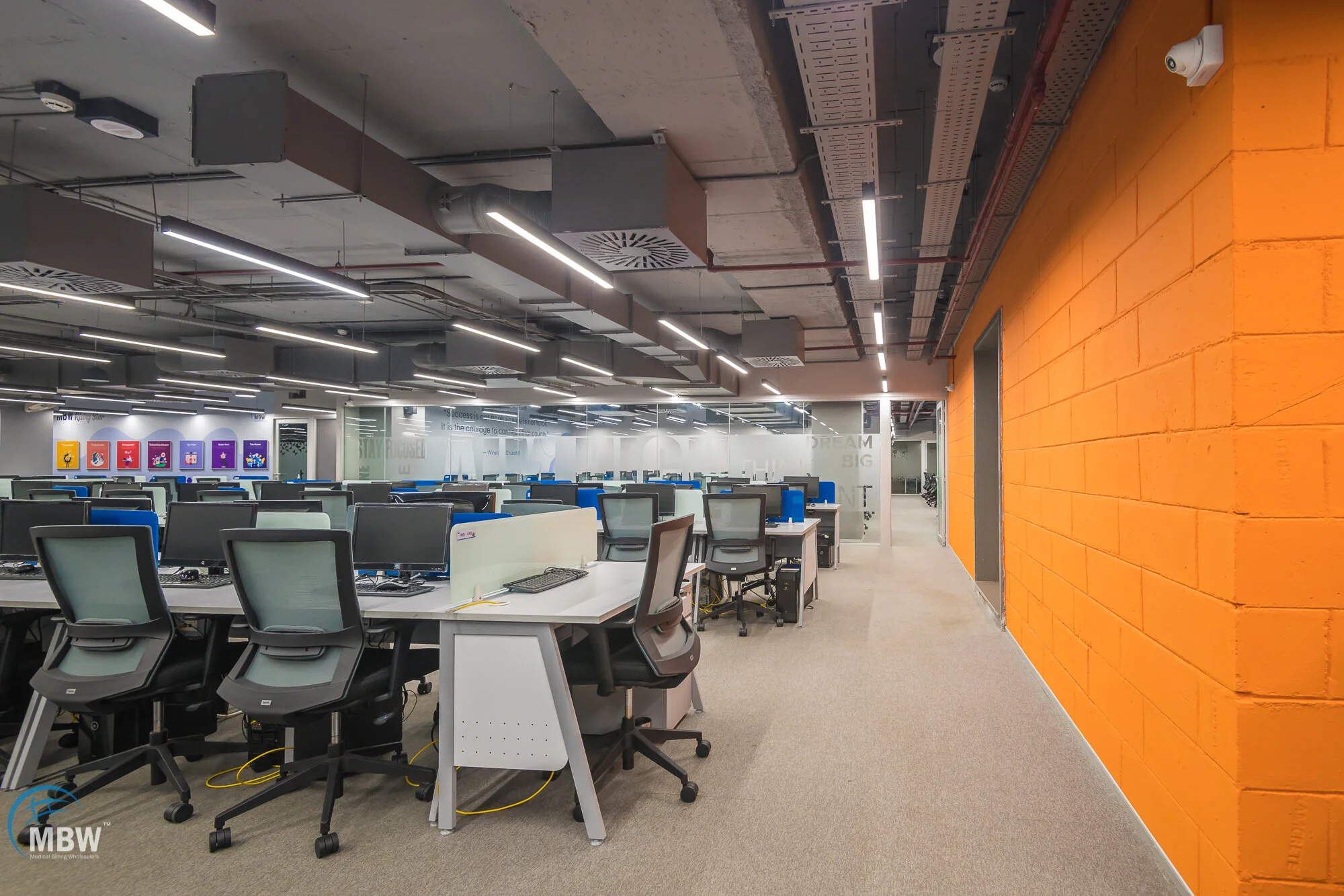 Open office with multiple desks, black and gray ergonomic chairs, and bright overhead lighting. An orange wall is on the right, and a glass partition separates the workspace from additional areas. There are colorful posters on the far wall.