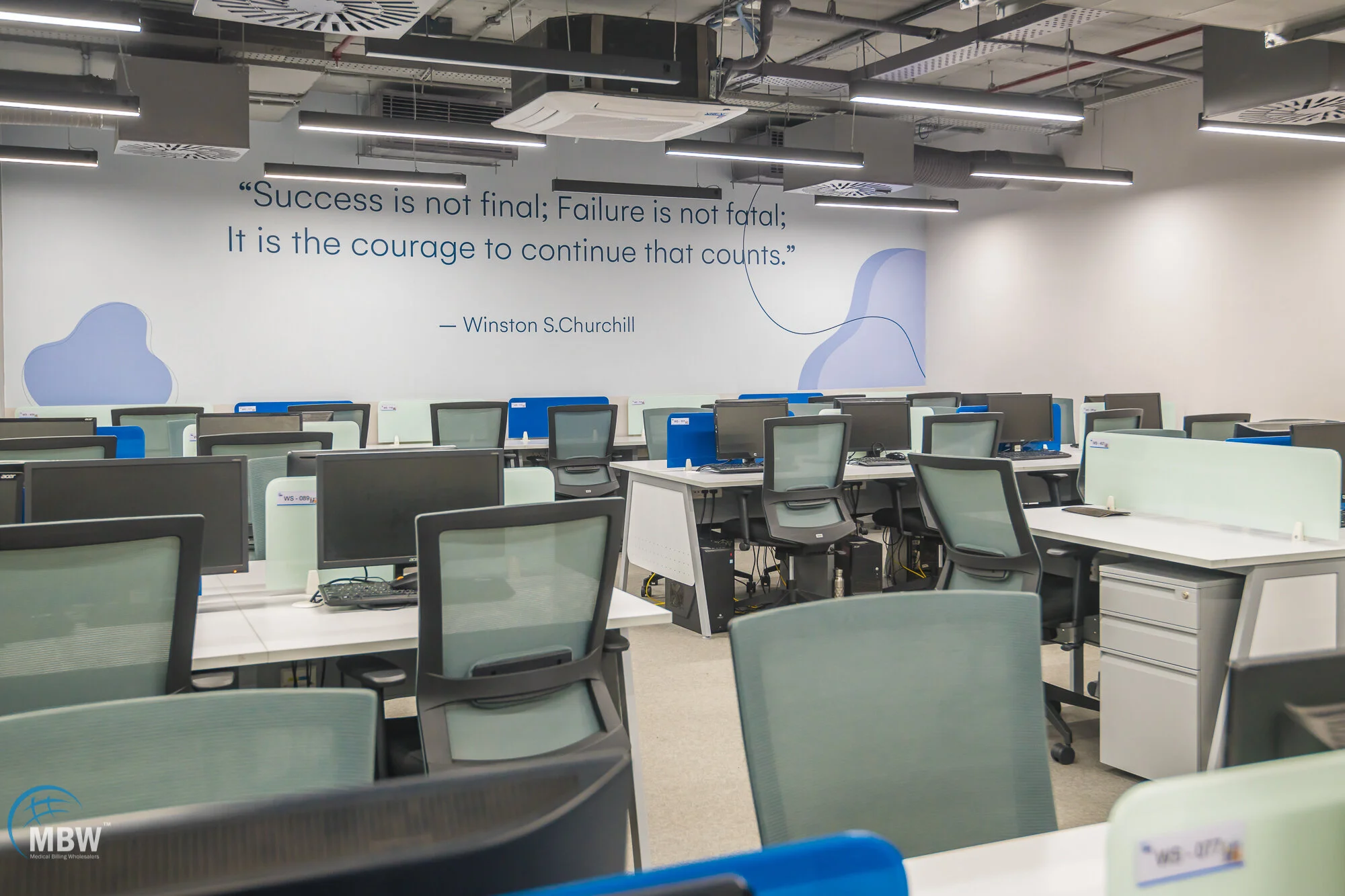 Empty office space with rows of desks and chairs, a quote on the wall by Winston S. Churchill, modern lighting, and ceiling vents.