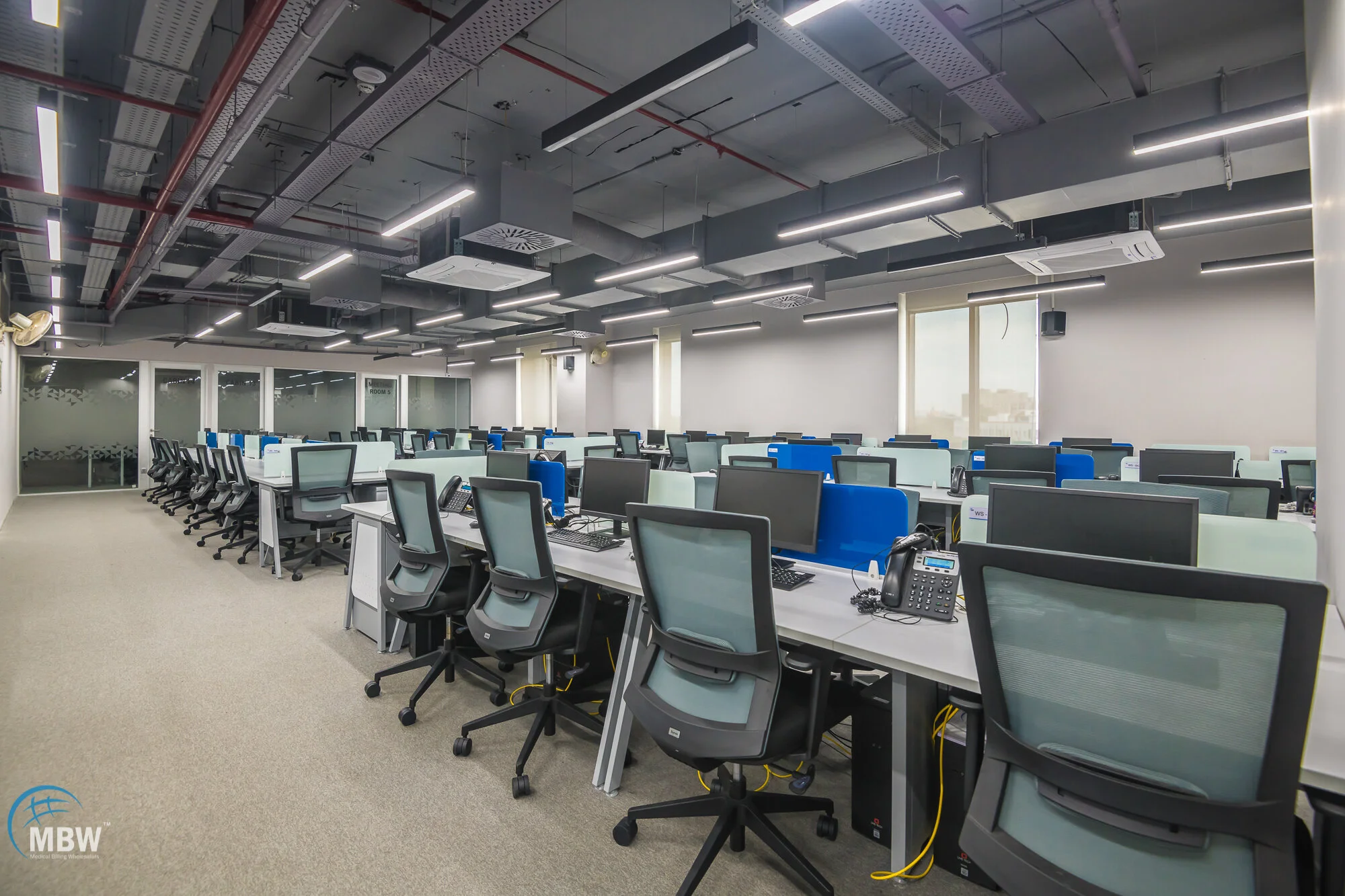 An empty modern office with rows of desks, black ergonomic chairs, computer monitors, phones, and cubicle dividers, with a ceiling featuring exposed ductwork and LED lighting.