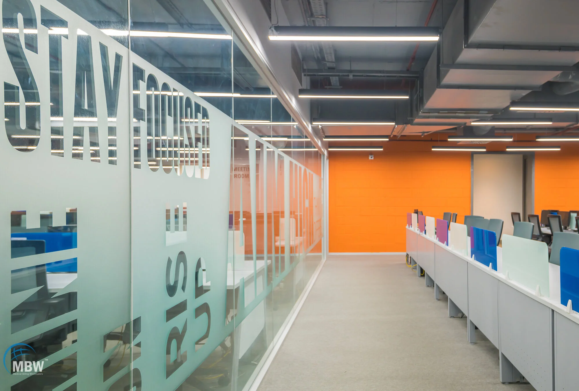 Empty office space with a row of desks and colorful dividers, glass partition with the words Bridal and Salon on it, orange wall at the back, modern lighting, and gray carpet.