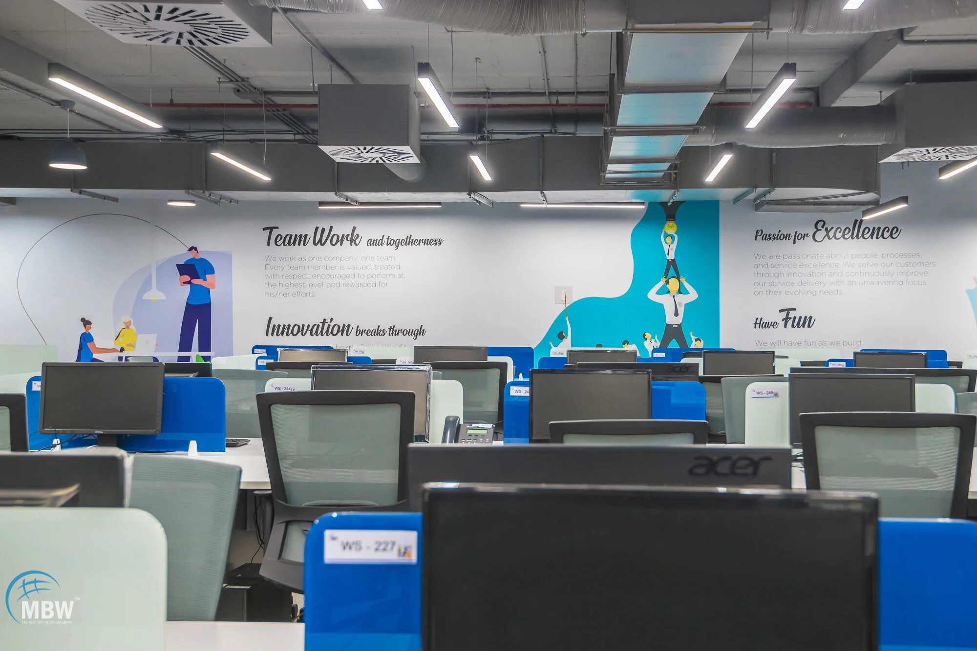 Modern office space with rows of desks and chairs, computer monitors, and a wall mural with motivational text and graphics related to teamwork, innovation, and excellence.