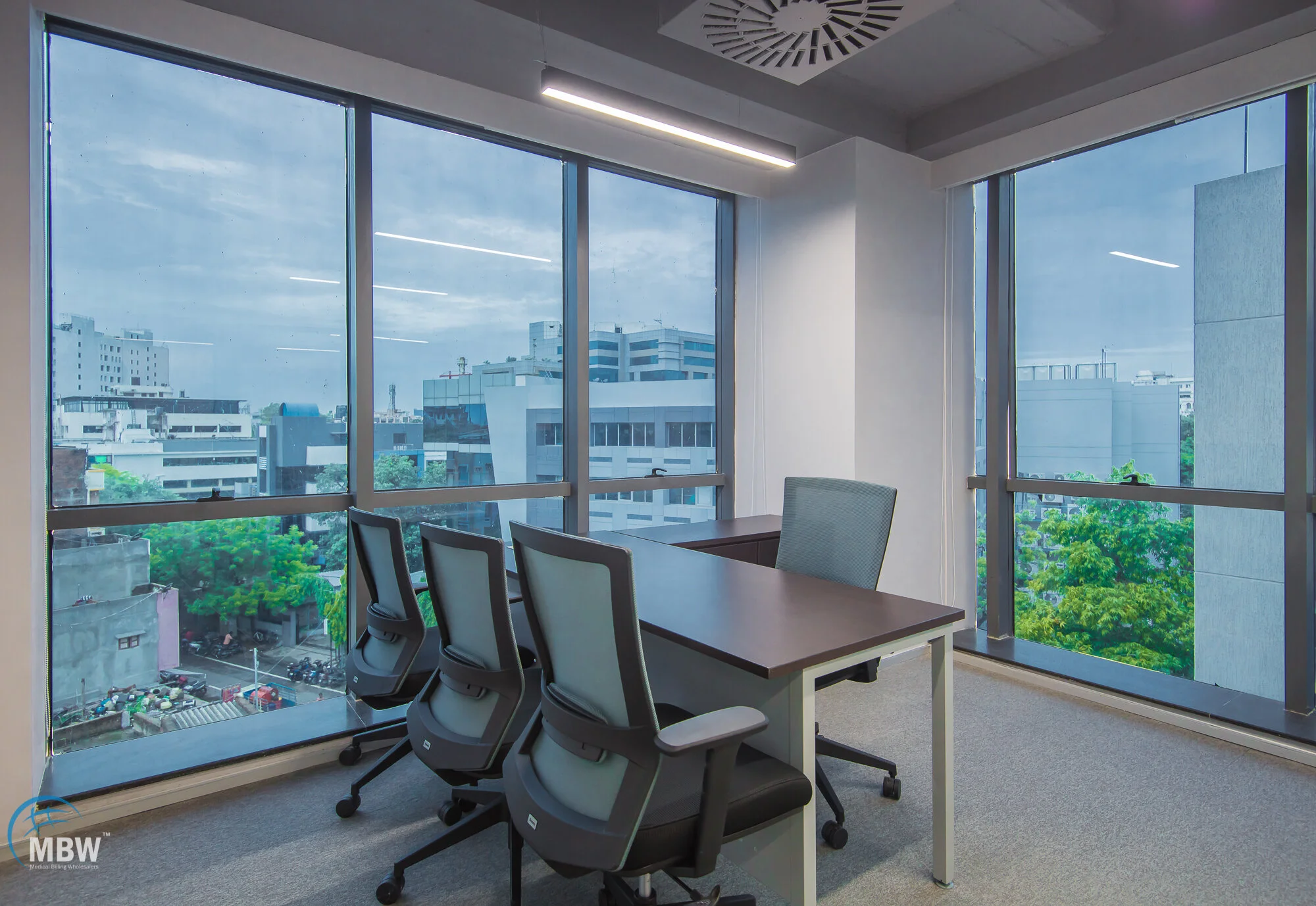 Modern office conference room with four black and gray chairs around a dark brown table, large windows offering a city view with buildings and greenery, white walls, and ceiling lights.