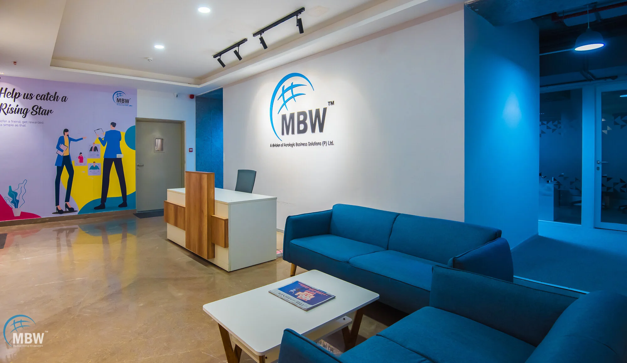 Office reception area with blue sofas, a white coffee table with magazines, a desk with a chair, a wall with the logo 'MBW', and a colorful mural on the left wall.