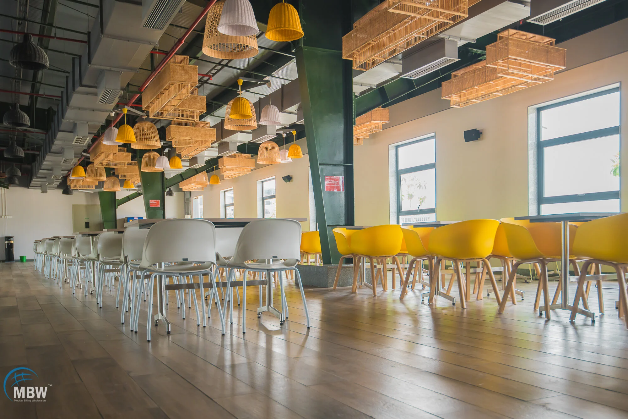 Empty restaurant with yellow chairs, white chairs, wooden flooring, and hanging wicker and yellow pendant lights.