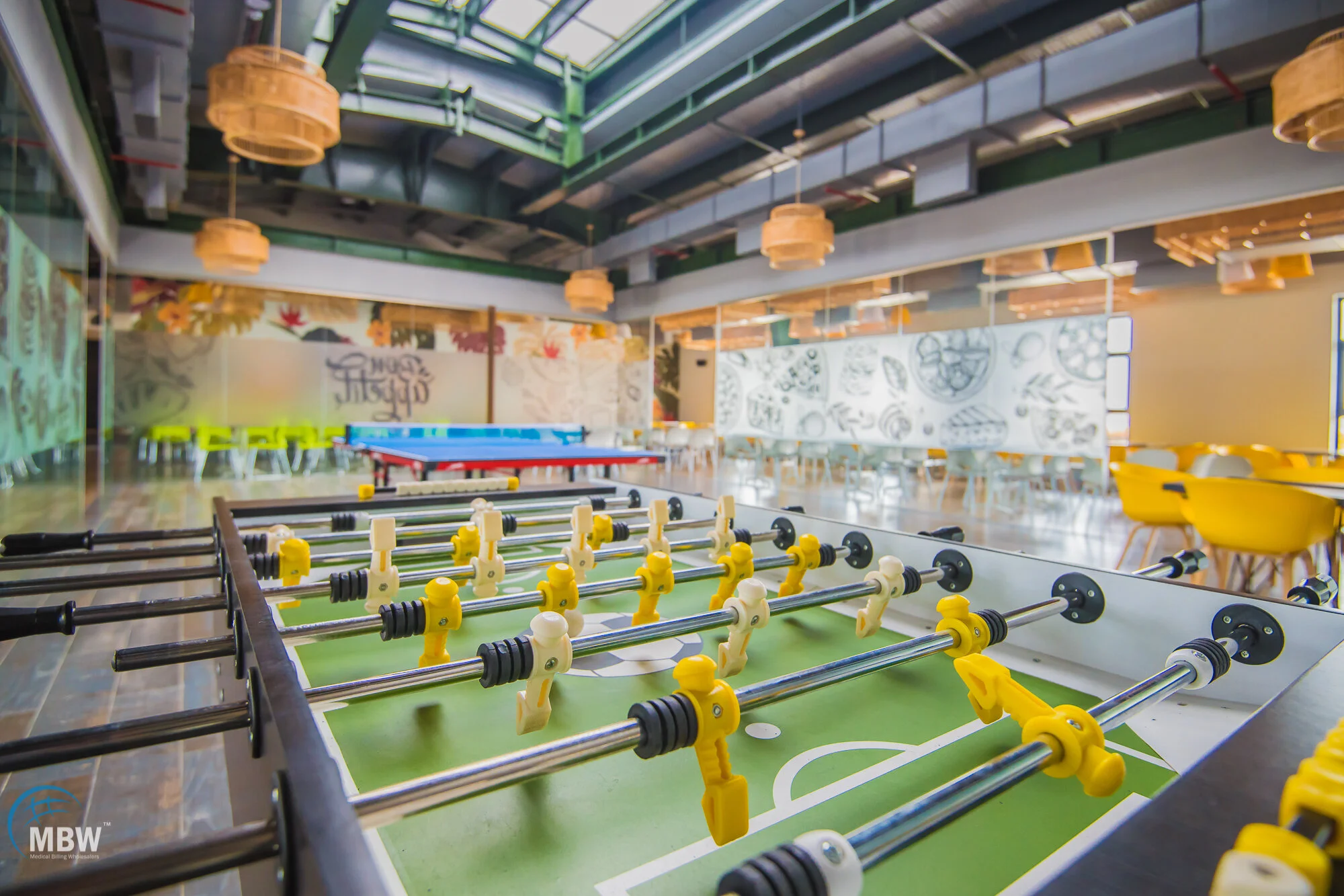 Indoor space with a foosball table in the foreground, seating area with yellow chairs on the right, and a colorful mural on the back wall. There are hanging wicker lamps and a glass ceiling.
