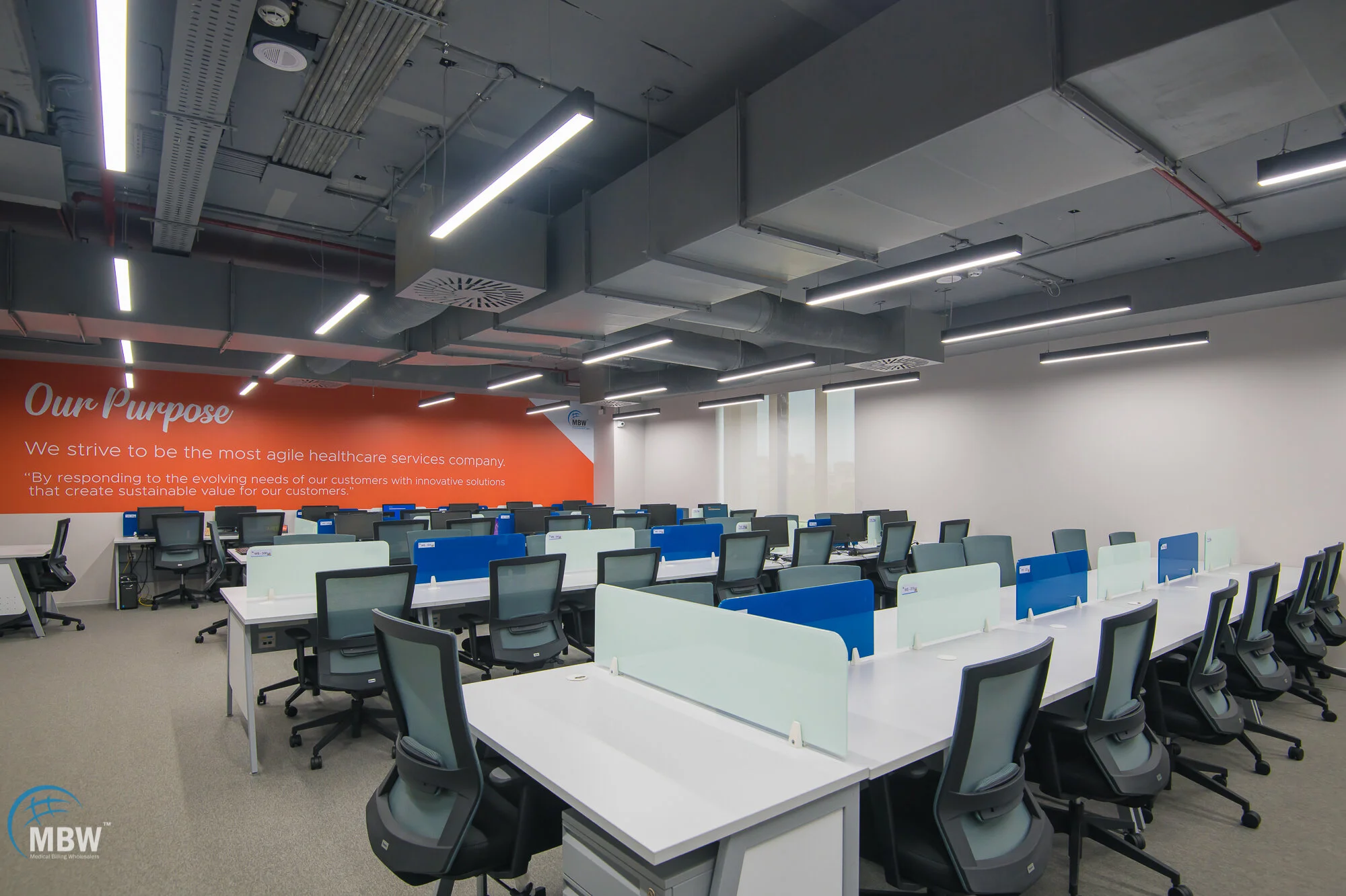 Empty modern office workspace with rows of desks, chairs, and privacy dividers, with a red wall displaying company values and mission statement.
