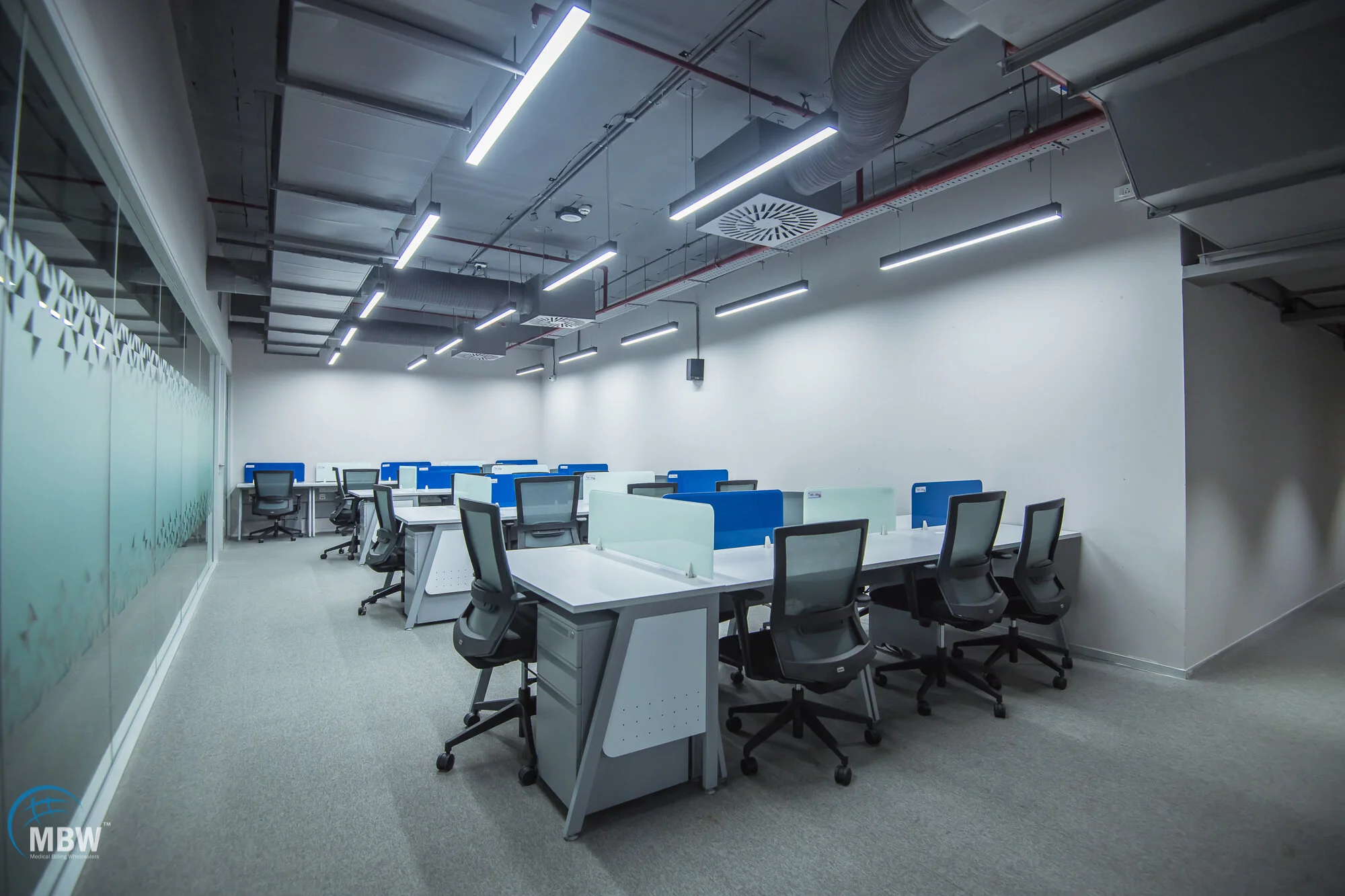 Empty modern office with desks, chairs, and partition screens, illuminated by ceiling lights.