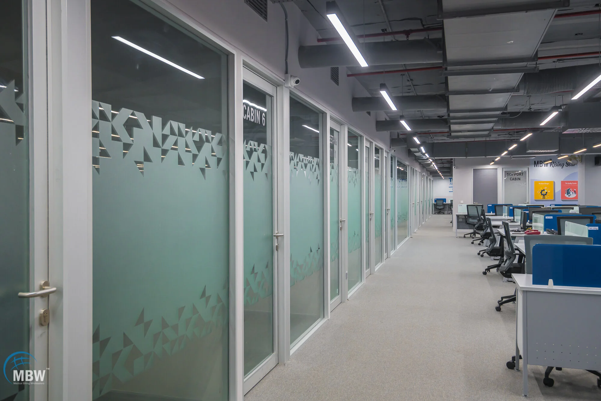 Empty office with glass-walled conference rooms on the left and workstations with chairs on the right, under bright ceiling lights.