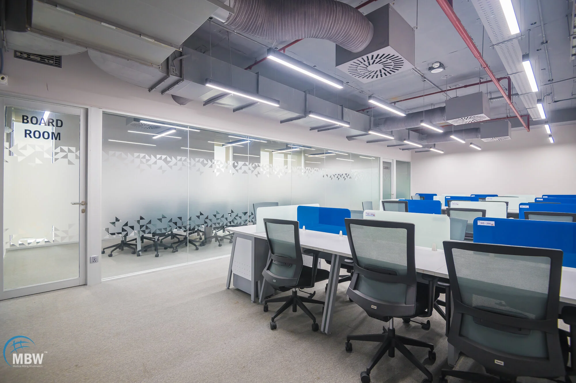 Modern office conference room with tables, chairs, and glass walls marked 'Board Room', ceiling with exposed ventilation and lighting fixtures.