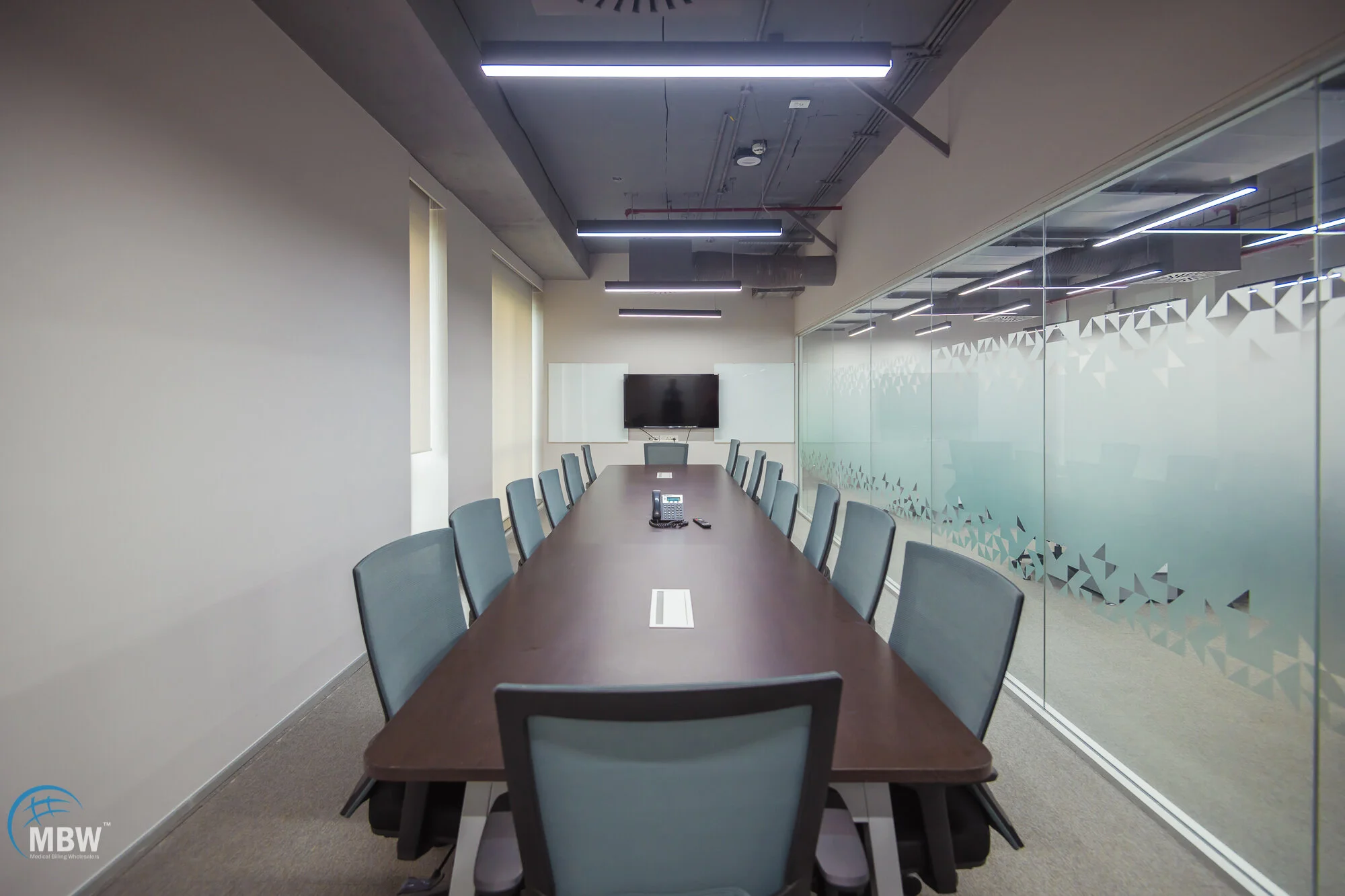 Empty conference room with a long wooden table, multiple gray office chairs, a conference phone, a wall-mounted TV, and frosted glass walls.